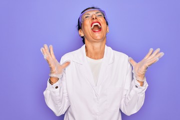 Naklejka premium Middle age senior scientist woman wearing coat and laboratory glasses over purple background crazy and mad shouting and yelling with aggressive expression and arms raised. Frustration concept.