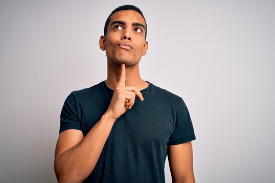 Young handsome african american man wearing casual t-shirt standing over white background Thinking concentrated about doubt with finger on chin and looking up wondering