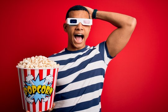 Young handsome african american man watching movie using 3d glasses eating popcorns Crazy and scared with hands on head, afraid and surprised of shock with open mouth