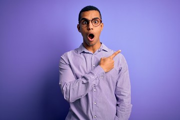 Handsome african american man wearing striped shirt and glasses over purple background Surprised pointing with finger to the side, open mouth amazed expression.