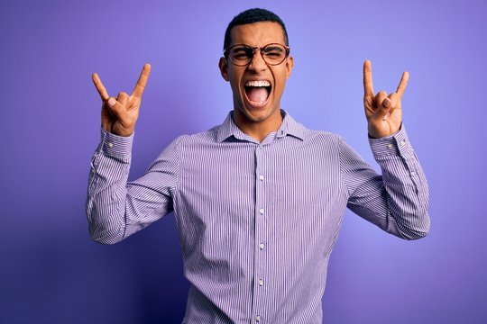 Handsome african american man wearing striped shirt and glasses over purple background shouting with crazy expression doing rock symbol with hands up. Music star. Heavy music concept.