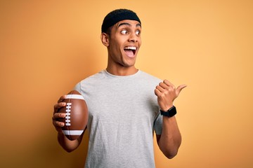 Young handsome african american man holding rugby ball over isolated yellow background pointing and showing with thumb up to the side with happy face smiling