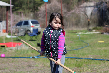 a little white dark-haired white girl with a ponytail outdoors holds a long wooden stick in her hands. Preschooler helps parents in the spring garden