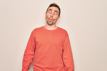 Young handsome man with blue eyes wearing casual sweater standing over white background making fish face with lips, crazy and comical gesture. Funny expression.