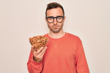 Handsome man with blue eyes holding bowl with healthy almonds snack over white background with a confident expression on smart face thinking serious