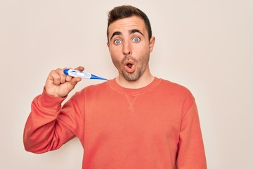 Young handsome man with blue eyes measuring temperature using digital thermometer scared and amazed...