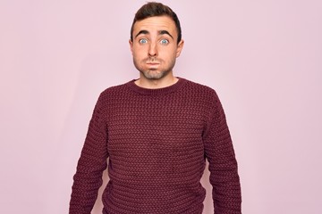 Young handsome man with blue eyes wearing casual sweater standing over pink background puffing cheeks with funny face. Mouth inflated with air, crazy expression.