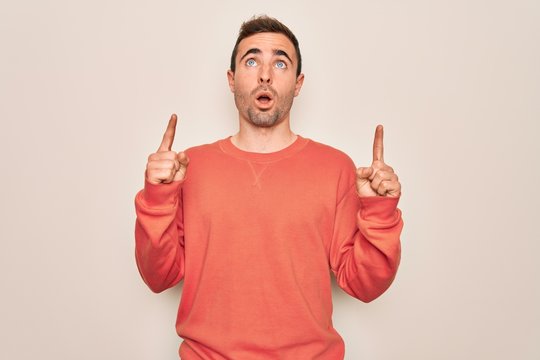Young handsome man with blue eyes wearing casual sweater standing over white background amazed and surprised looking up and pointing with fingers and raised arms.