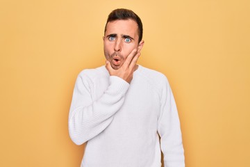 Young handsome man with blue eyes wearing casual sweater standing over yellow background Looking fascinated with disbelief, surprise and amazed expression with hands on chin