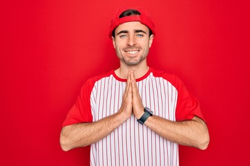 Young handsome sporty man with blue eyes wearing striped baseball t-shirt and cap praying with hands together asking for forgiveness smiling confident.