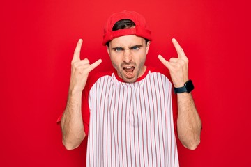 Young handsome sporty man with blue eyes wearing striped baseball t-shirt and cap shouting with crazy expression doing rock symbol with hands up. Music star. Heavy concept.
