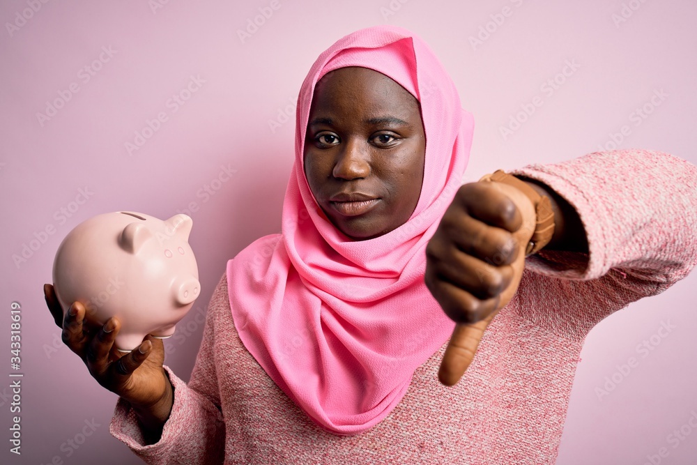 Poster young african american plus size woman wearing muslim hijab holding pink piggy bank with angry face,