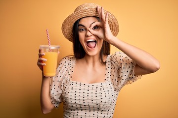 Young beautiful brunette woman on vacation wearing summer hat drinking orange juice with happy face smiling doing ok sign with hand on eye looking through fingers