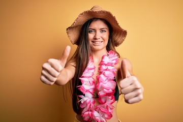 Young beautiful woman with blue eyes on vacation wearing bikini and hawaiian lei approving doing positive gesture with hand, thumbs up smiling and happy for success. Winner gesture.