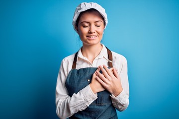 Young beautiful baker woman with blue eyes wearing apron and cap over blue background smiling with hands on chest with closed eyes and grateful gesture on face. Health concept.