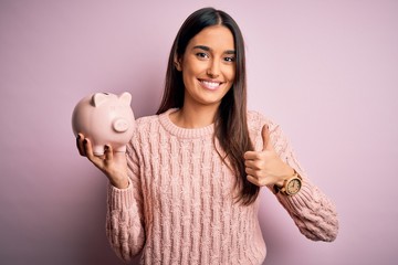 Young beautiful brunette woman holding piggy bank saving money for retirement happy with big smile...