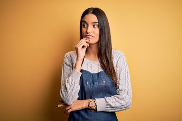 Young beautiful baker woman wearing apron uniform cooking over yellow background with hand on chin thinking about question, pensive expression. Smiling with thoughtful face. Doubt concept.