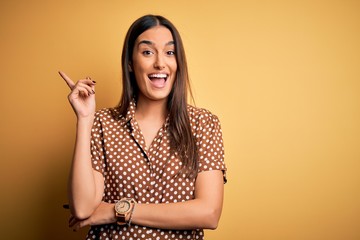 Young beautiful brunette woman wearing casual shirt over isolated yellow background with a big smile on face, pointing with hand and finger to the side looking at the camera.