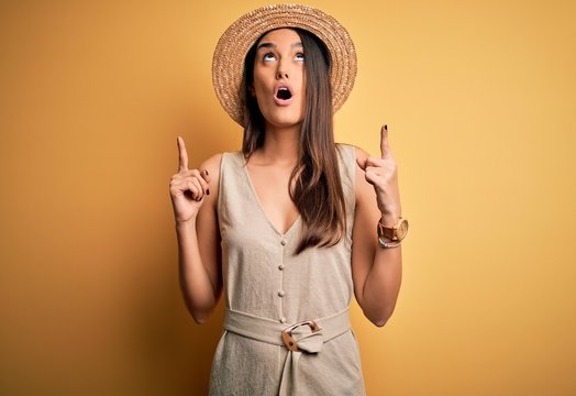 Young beautiful brunette woman on vacation wearing casual dress and hat amazed and surprised looking up and pointing with fingers and raised arms.