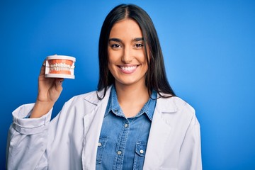 Young beautiful brunette dentist woman wearing coat holding plastic teeth prosthesis with a happy face standing and smiling with a confident smile showing teeth