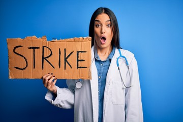 Young doctor woman wearing stethoscope holding cardboard banner protesting in strike scared in shock with a surprise face, afraid and excited with fear expression