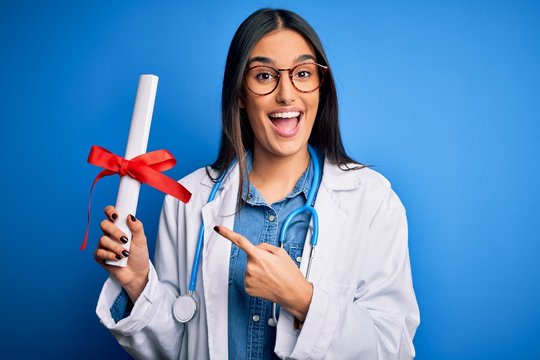 Young beautiful brunette doctor woman wearing glasses and coat holding diploma degree very happy pointing with hand and finger