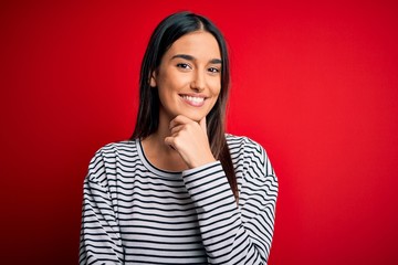 Young beautiful brunette woman wearing casual striped t-shirt over red background looking confident at the camera smiling with crossed arms and hand raised on chin. Thinking positive.