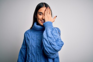 Young beautiful brunette woman wearing casual turtleneck sweater over white background covering one eye with hand, confident smile on face and surprise emotion.