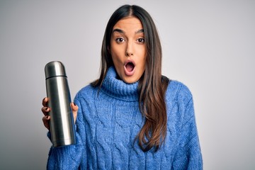 Young beautiful brunette tourist woman holding thermo with water over white background scared in...