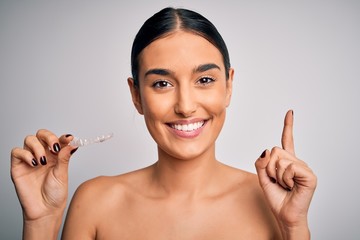 Young beautiful brunette woman holding dental aligner orthodontic to teeth corretion surprised with...