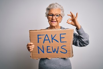 Senior grey-haired woman holding banner with fake news message over white background doing ok sign...