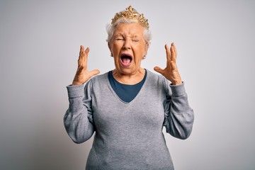 Senior beautiful grey-haired woman wearing golden queen crown over white background celebrating mad...