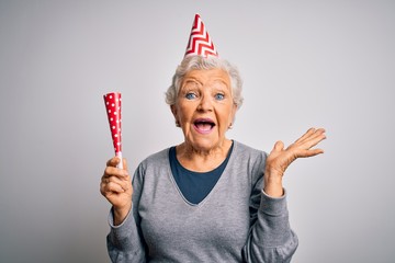 Senior beautiful grey-haired woman celebrating birthday wearing funny hat holding trumpet very...