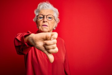 Senior beautiful grey-haired woman wearing casual shirt and glasses over red background looking unhappy and angry showing rejection and negative with thumbs down gesture. Bad expression.