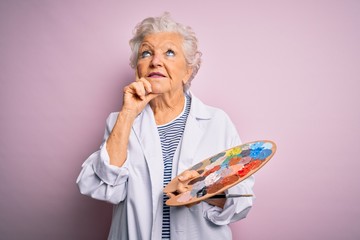 Senior beautiful grey-haired artist woman painting using brush and palette over pink background...
