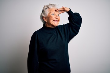 Senior beautiful woman wearing casual black sweater standing over isolated white background very happy and smiling looking far away with hand over head. Searching concept.