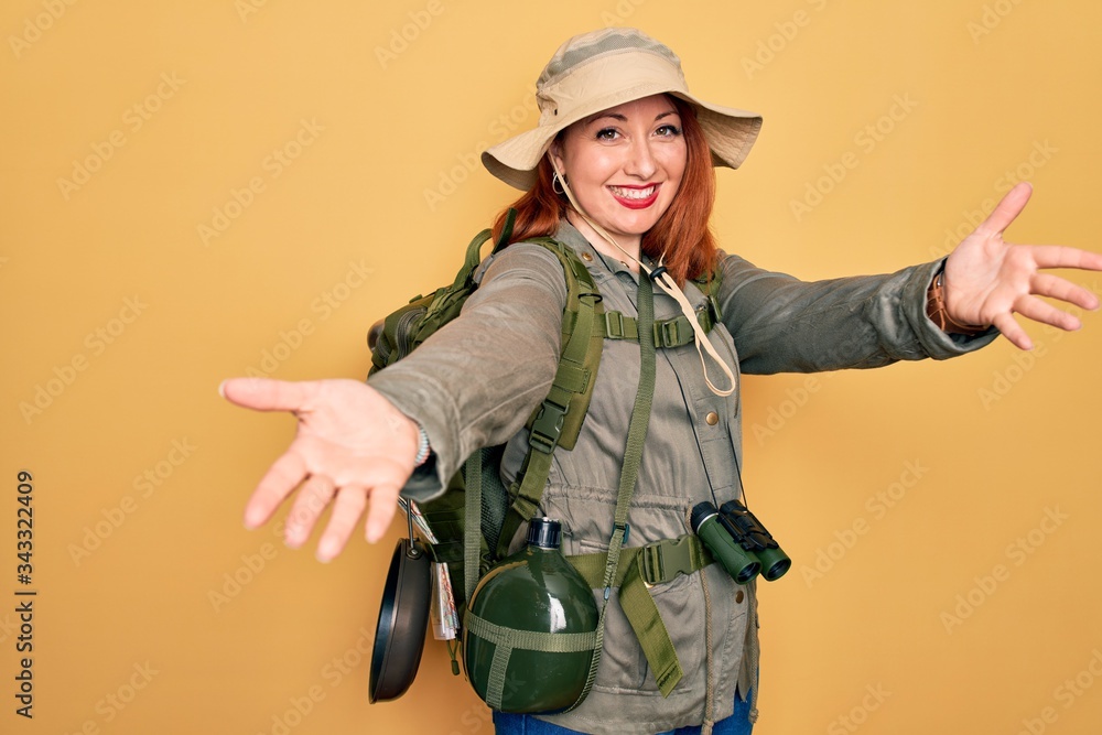 Sticker young redhead backpacker woman hiking wearing backpack and hat over yellow background looking at the