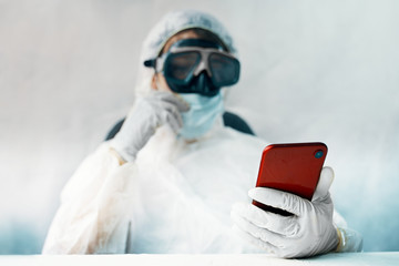 Doctor in the protective suit is online. Virologist holds smartphone in gloves. communication in...