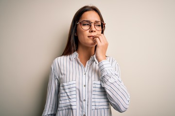 Young beautiful brunette woman wearing casual shirt and glasses over white background looking stressed and nervous with hands on mouth biting nails. Anxiety problem.