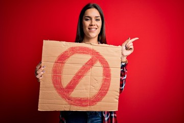 Young beautiful brunette woman holding banner with prohibited signal over red background very happy...