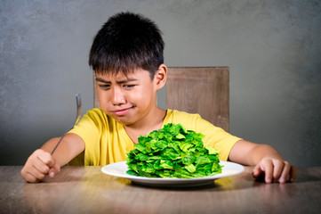 upset and disgusted hispanic kid sitting on table in front of spinach plate unhappy rejecting the...