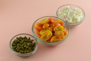 Fresh raw grape tomatoes, chopped shallot onions and capers in glass ingredient bowls on pink background