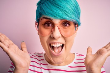 Young beautiful woman with blue fashion hair wearing fanny glasses with hearts very happy and excited, winner expression celebrating victory screaming with big smile and raised hands