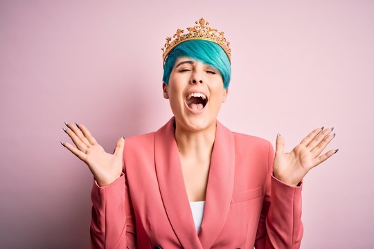 Young business woman with blue fashion hair wearing queen crown over pink isolated background celebrating mad and crazy for success with arms raised and closed eyes screaming excited. Winner concept