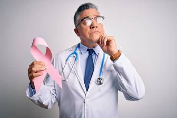 Middle age handsome grey-haired doctor man holding pink cancer ribbon serious face thinking about...