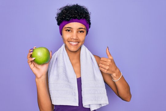 Beautiful african american afro sporty woman doing exercise wearing towel eating green apple happy with big smile doing ok sign, thumb up with fingers, excellent sign