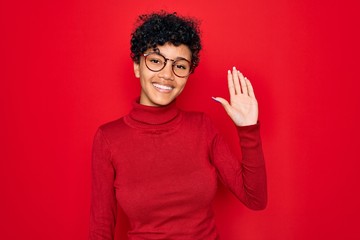 Young beautiful african american afro woman wearing turtleneck sweater and glasses Waiving saying...