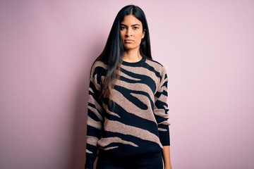 Young beautiful hispanic woman wearing animal print sweater over pink background with serious expression on face. Simple and natural looking at the camera.