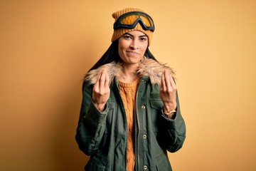 Young beautiful hispanic woman wearing ski glasses and coat for winter weather doing money gesture with hands, asking for salary payment, millionaire business