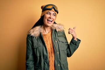 Young beautiful hispanic woman wearing ski glasses and coat for winter weather smiling with happy face looking and pointing to the side with thumb up.